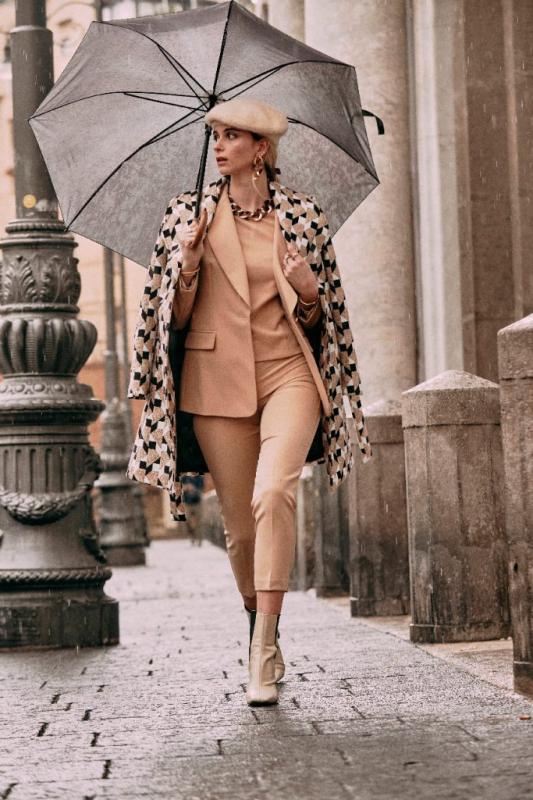 modella passeggia con l'ombrello e indossa un cappotto a fantasia geometrica sopra un tailleur beige